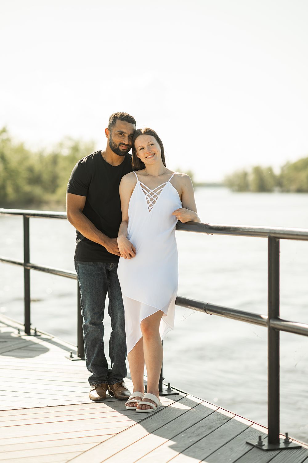 A couple poses together on a wooden deck overlooking a waterfront during a sunny day.
