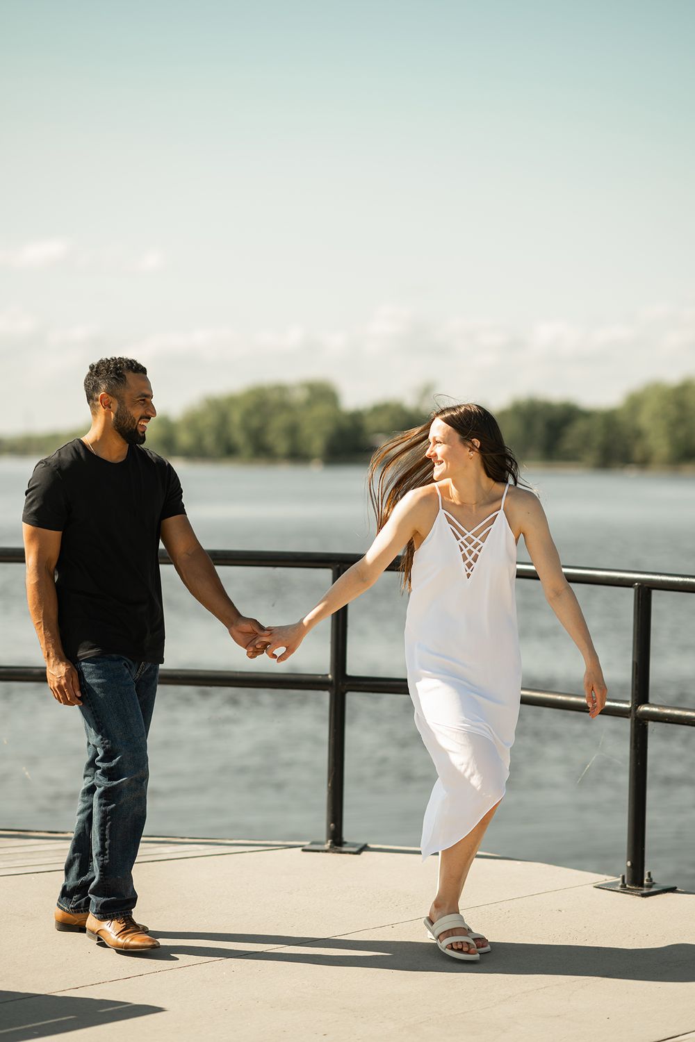 A couple in black and white outfits hold hands while walking along a waterfront railing on a sunny day.