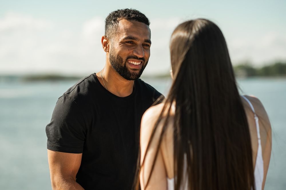 A couple shares a romantic moment by the water's edge, laughing together near a metal railing.
