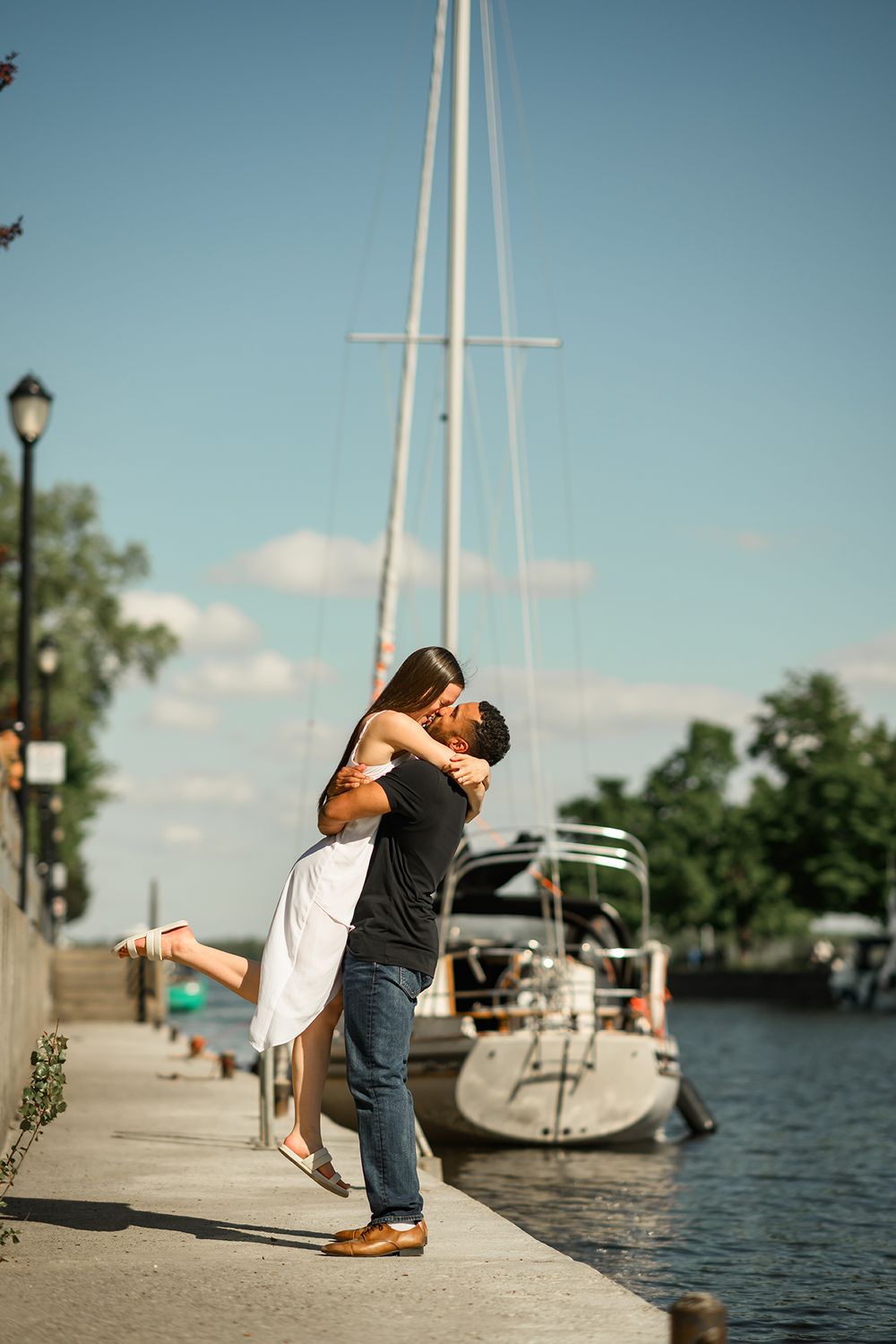 A couple embraces and dances playfully on a marina dock with sailboats and water in the background.