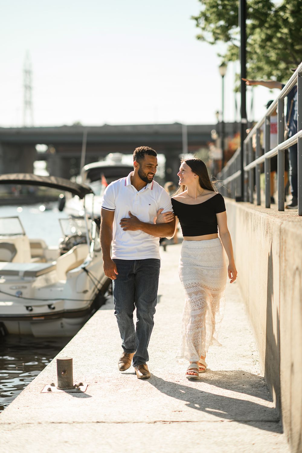 A couple walks playfully along a marina walkway with boats docked in the background.