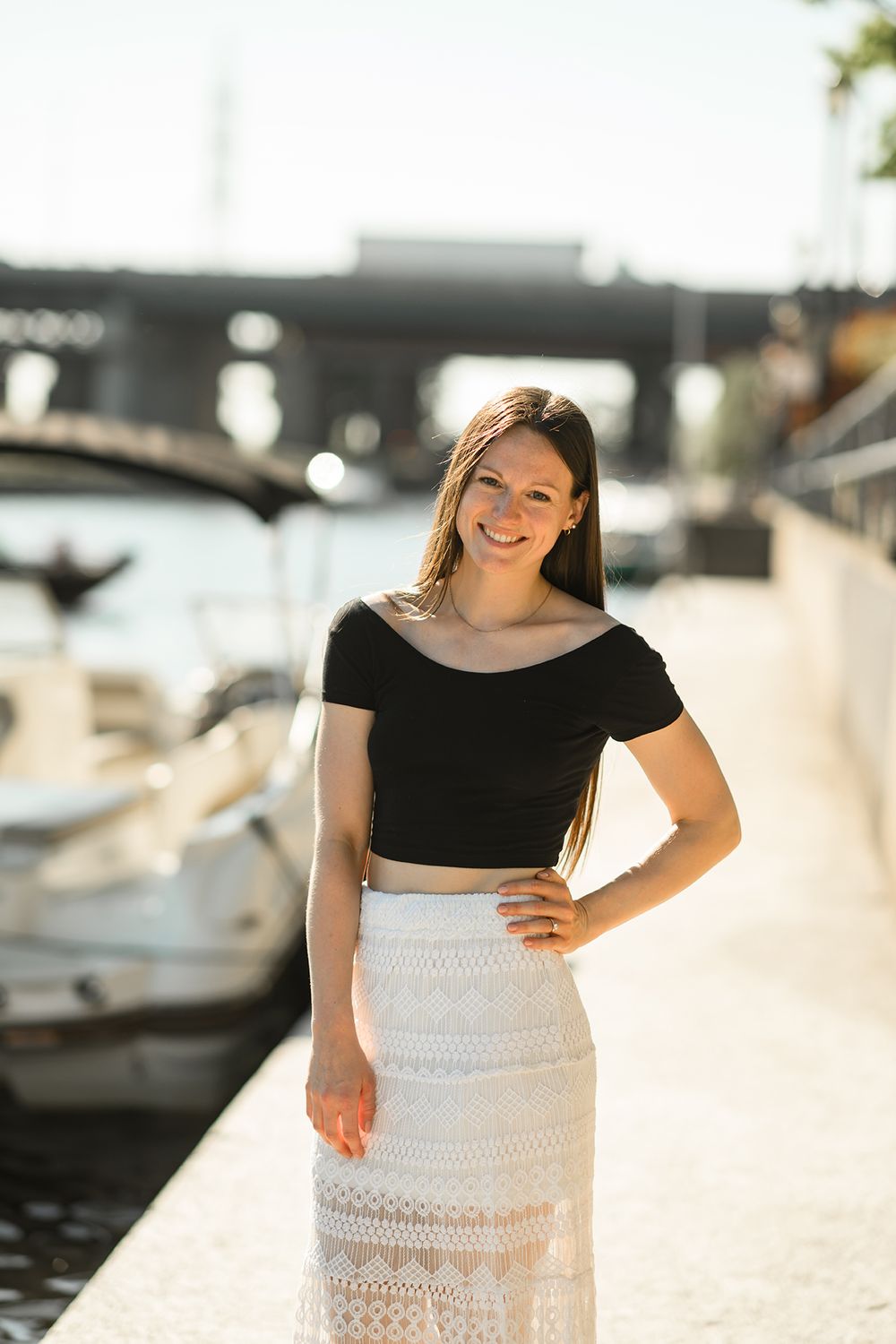 Woman in black top and white lace skirt poses by marina dock in bright sunlight.