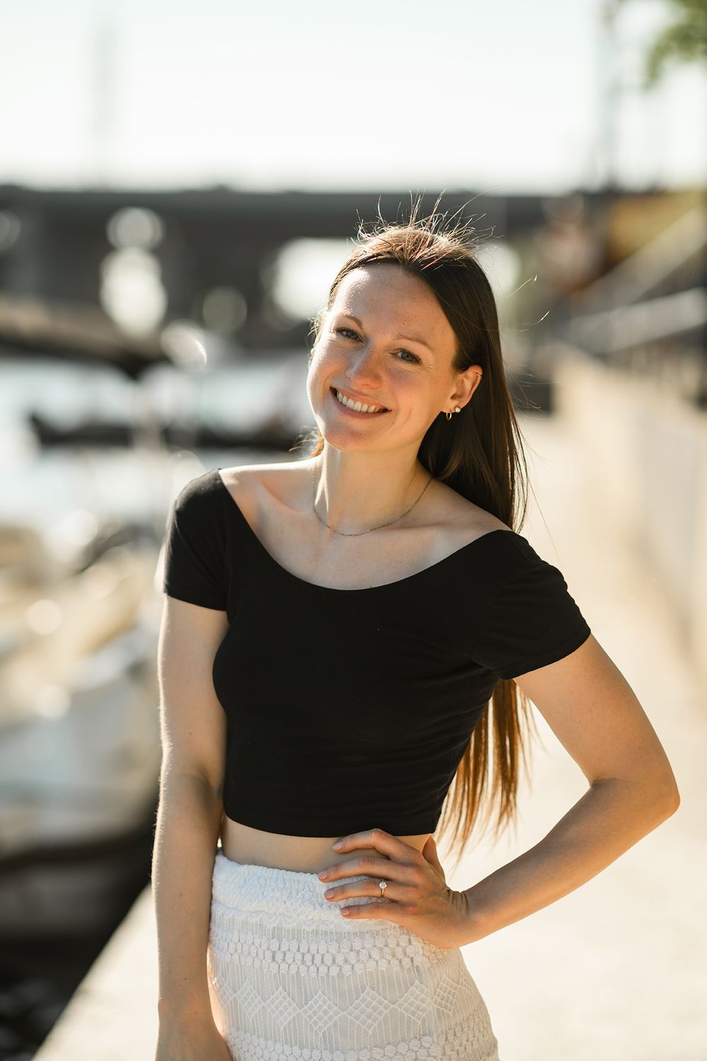 Woman in black crop top and white skirt smiling outdoors at marina during golden hour.