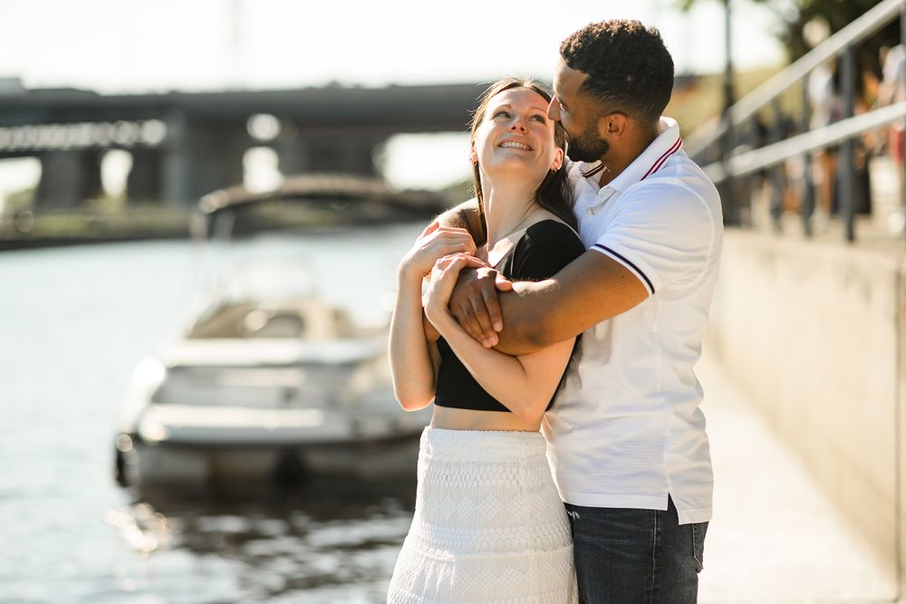 A joyful couple laughing and embracing by the waterfront with a boat and bridge in the background.