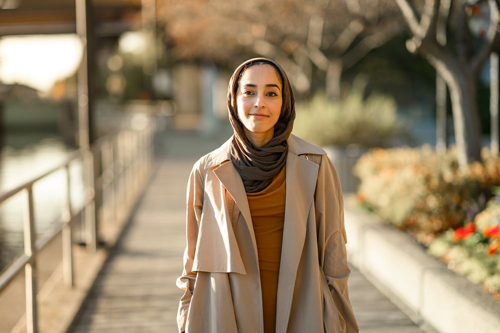 A person wearing a beige coat and brown turtleneck stands outdoors in autumn sunlight.