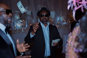 Money rains down as people celebrate at a dimly lit nightclub party with pink lighting effects.