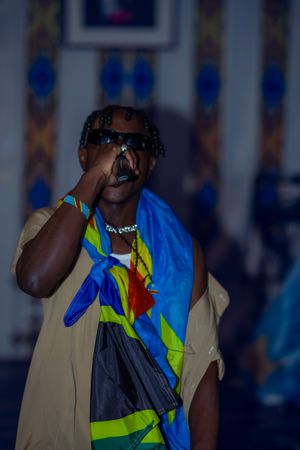 A performer in a bright blue and yellow outfit with sunglasses sings into a microphone on stage during a show.