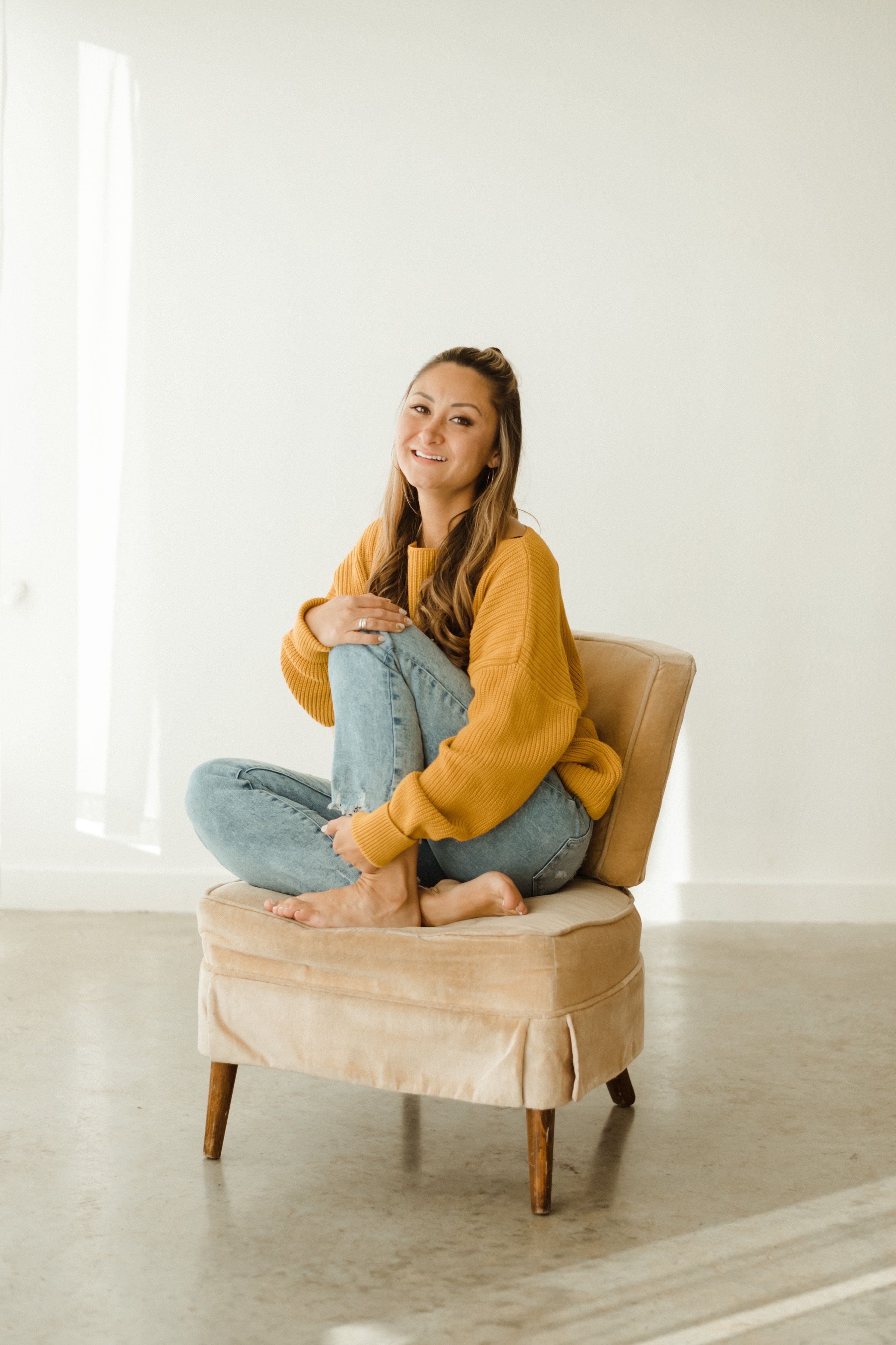 Person in mustard cardigan and jeans relaxing on beige upholstered chair in minimal white room setting.