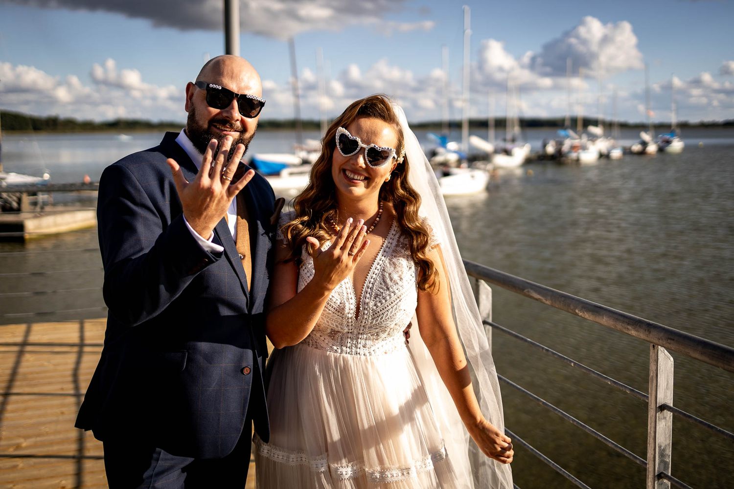 Newlyweds showing off rings on a marina dock with boats in background.