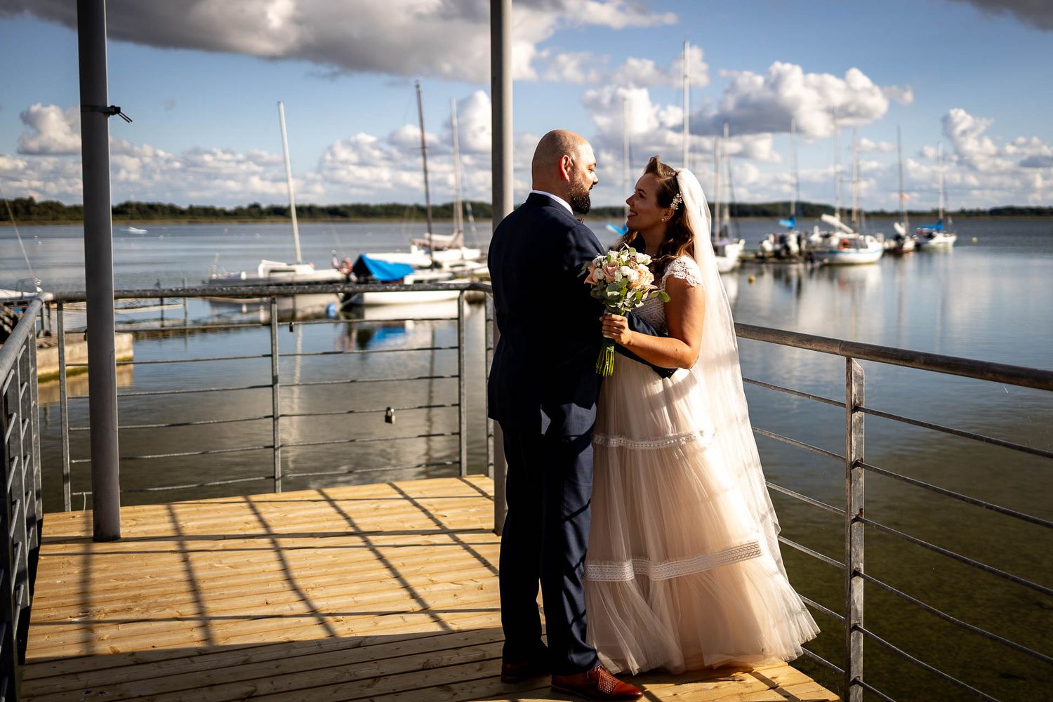 Couple in wedding attire standing on dock with marina and boats in background.