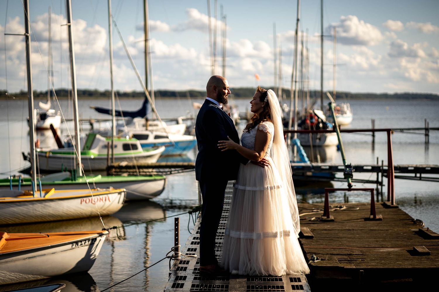 Couple standing on marina dock with sailboats and water in background during sunset.