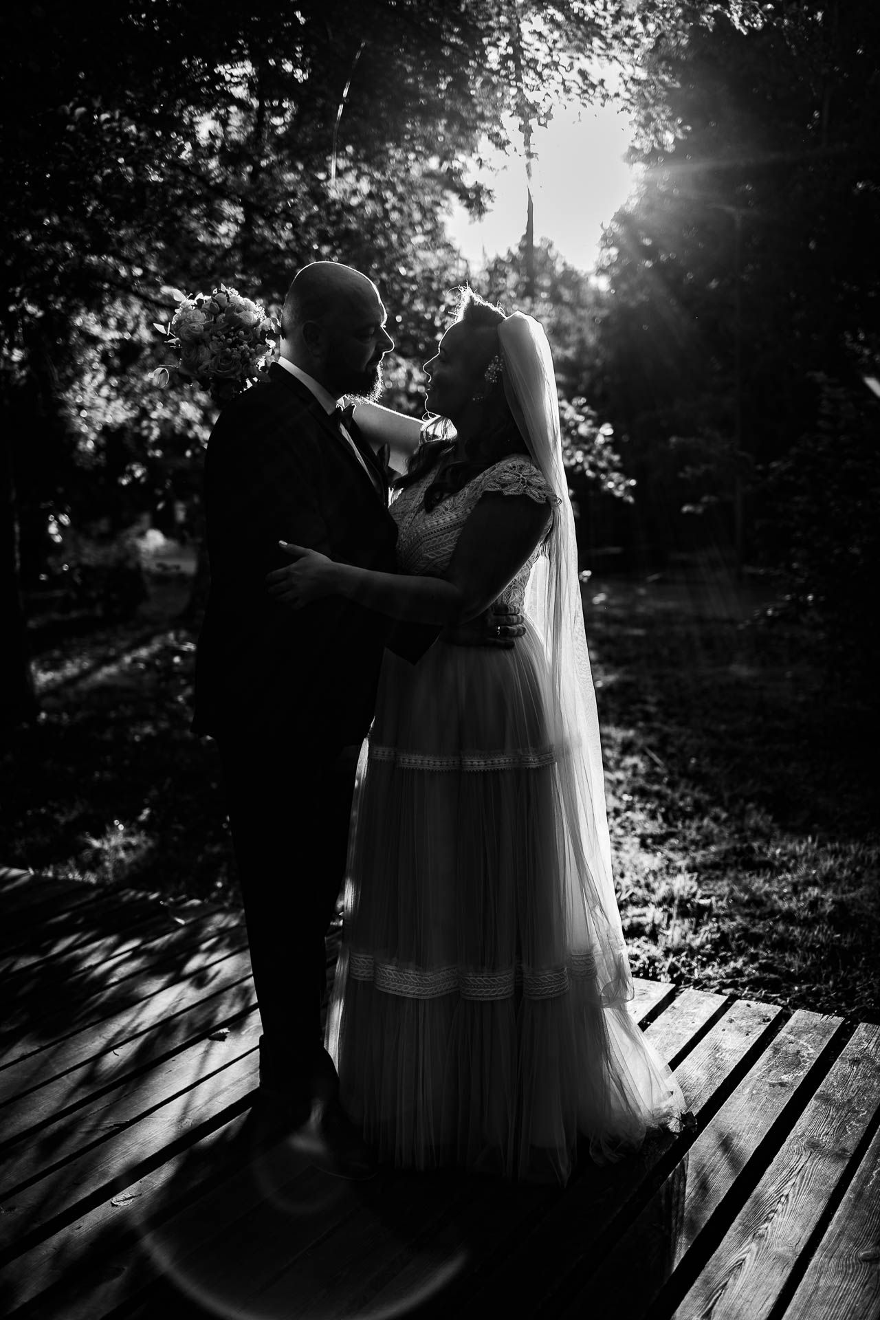 Silhouetted wedding couple on wooden deck with sunlight streaming through trees.