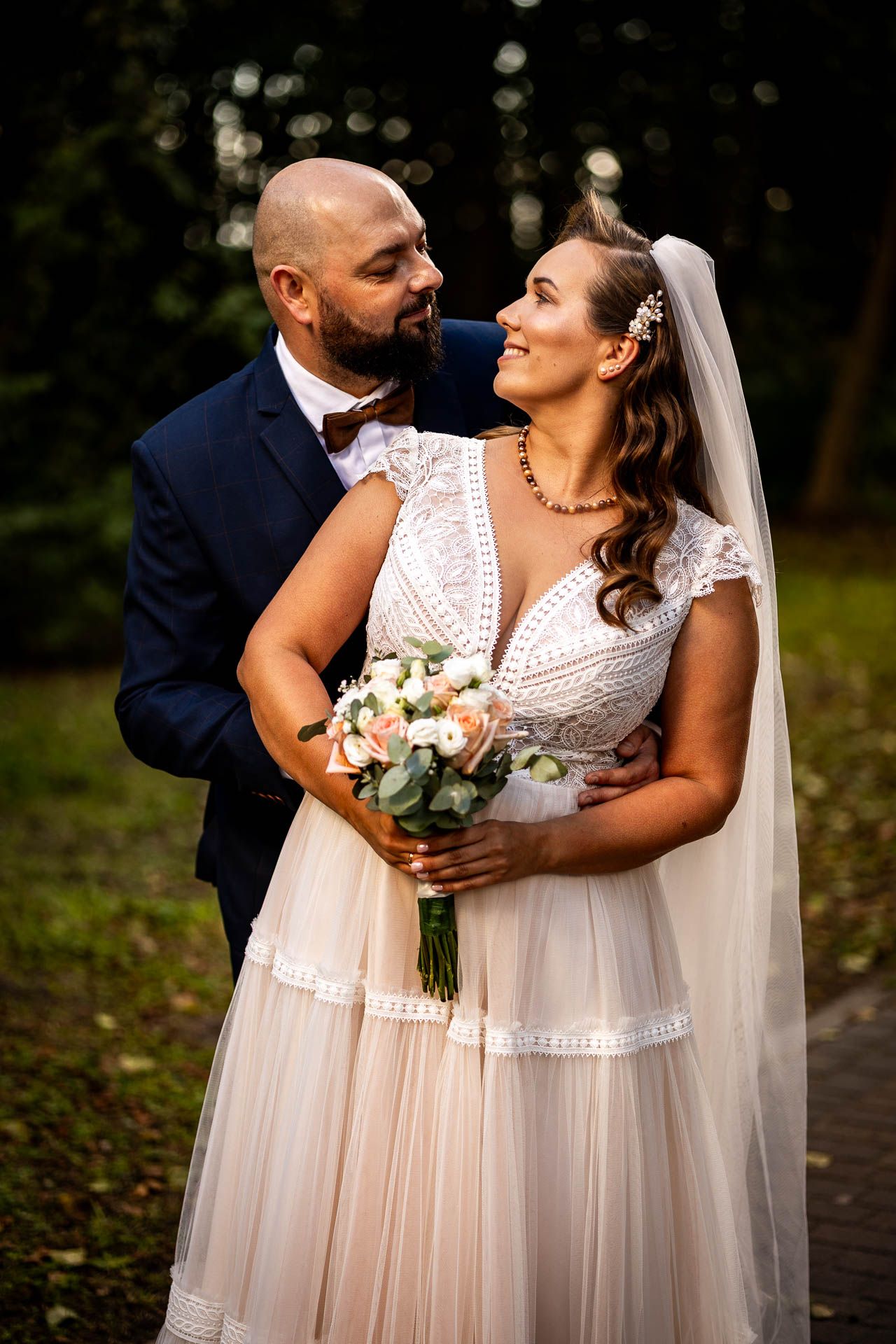 Wedding couple embracing outdoors while holding a white and peach floral bouquet.