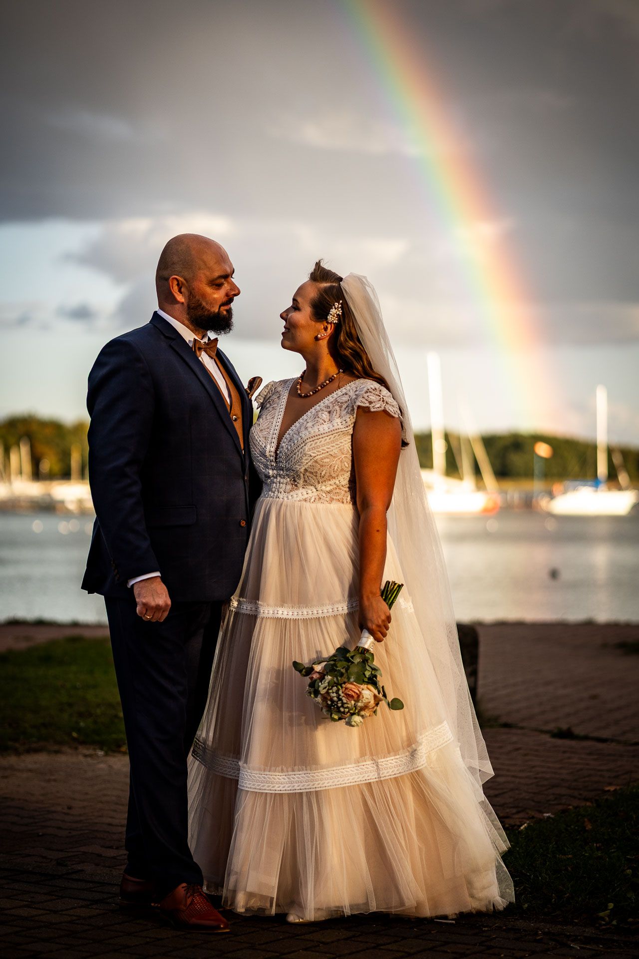 Wedding couple poses against waterfront with stunning rainbow in background at sunset.