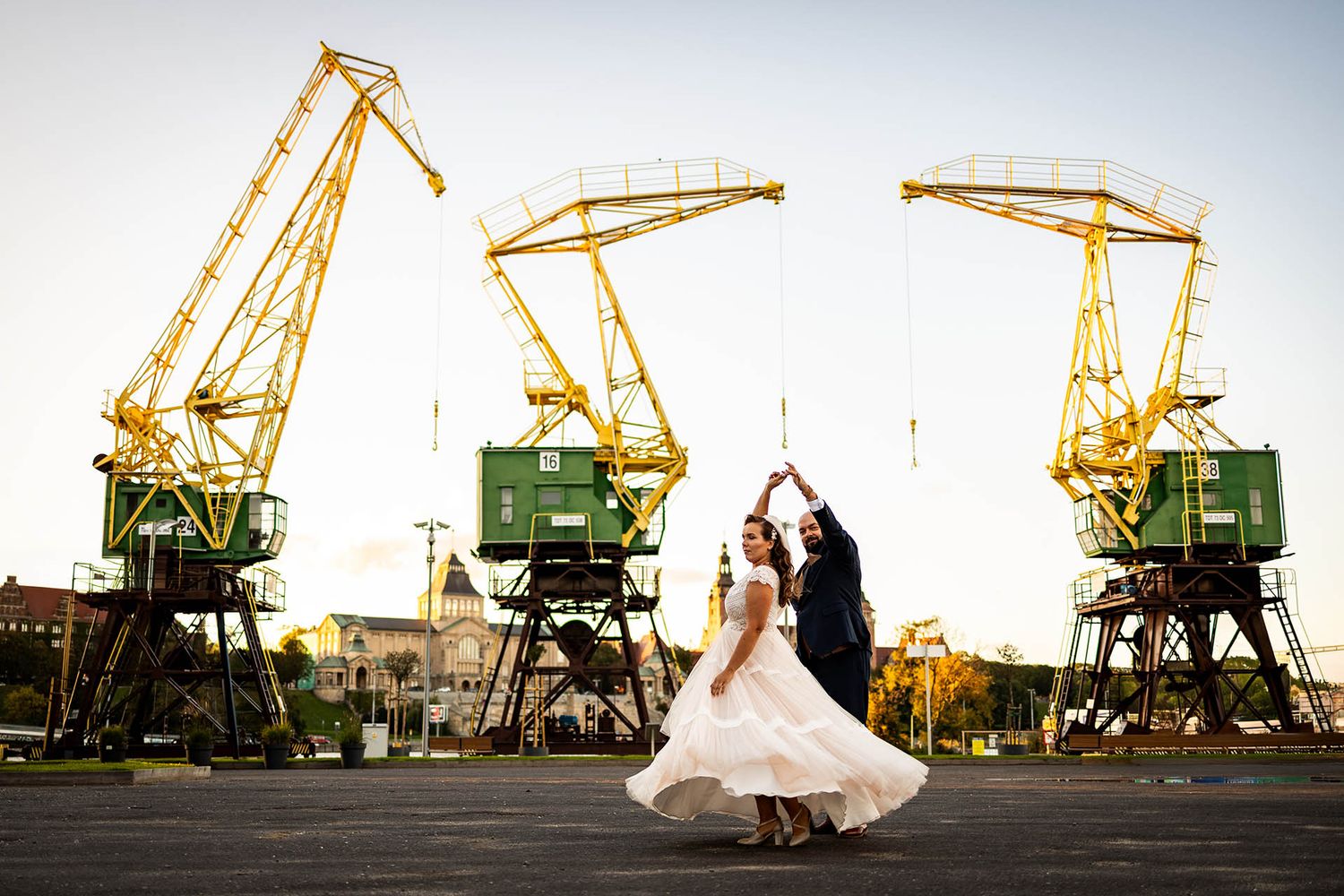 Wedding couple poses dramatically against industrial cranes and sunset at a port location.
