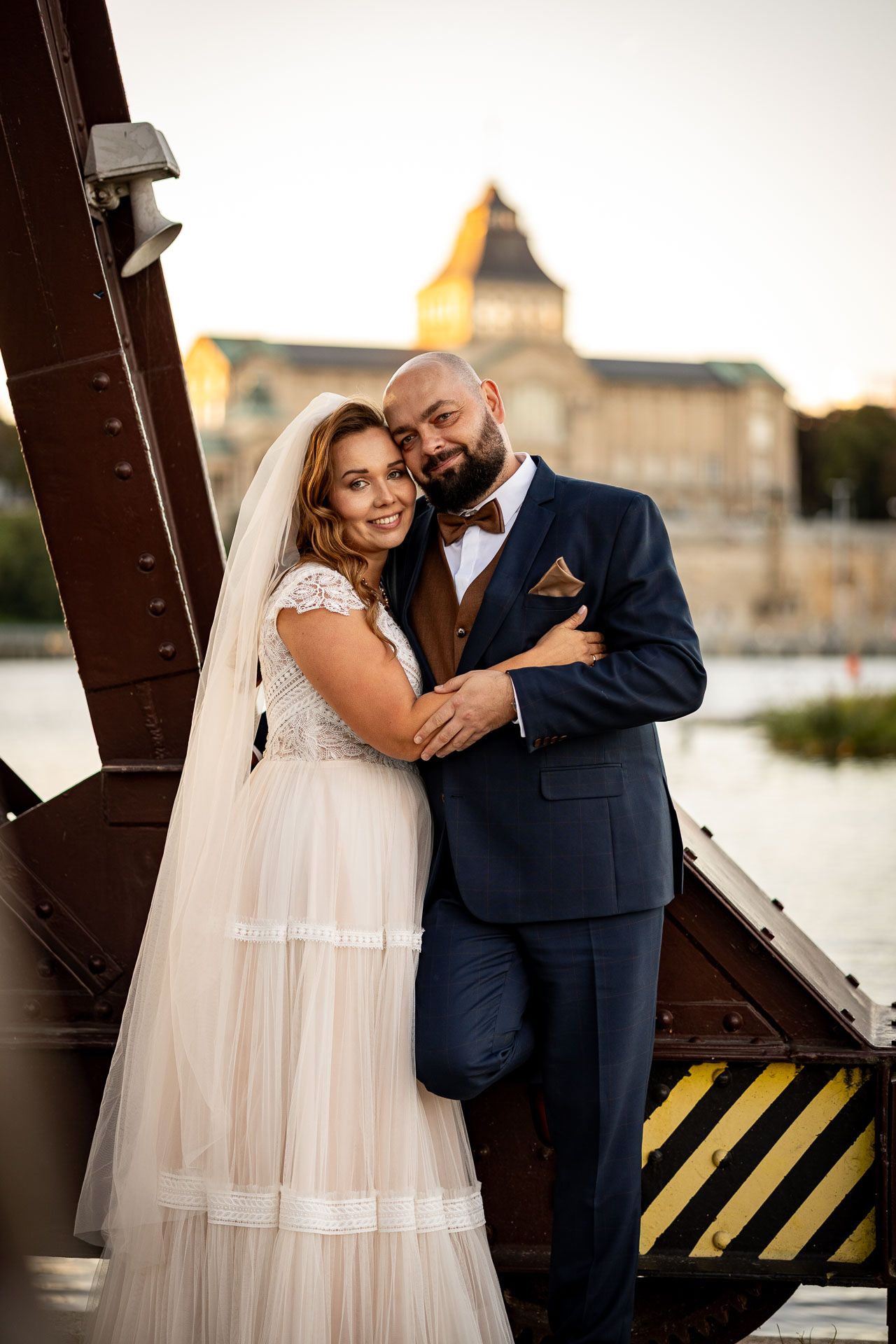 A romantic wedding couple embraces against an industrial backdrop with metal beams at sunset.
