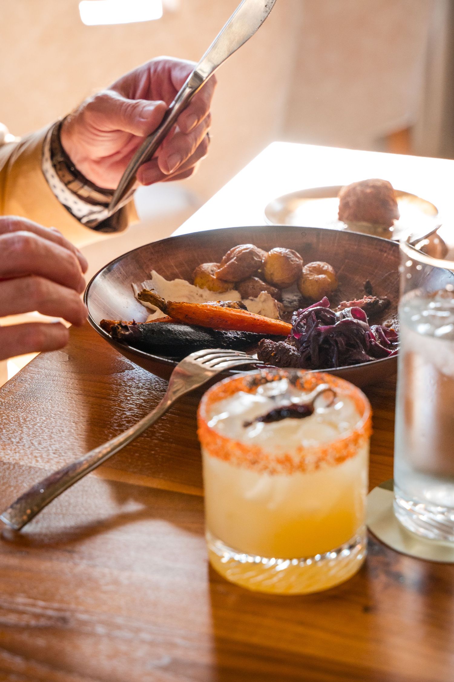 Hands serving braised meat and vegetables in a pan alongside a citrus cocktail with salt rim on wooden table.