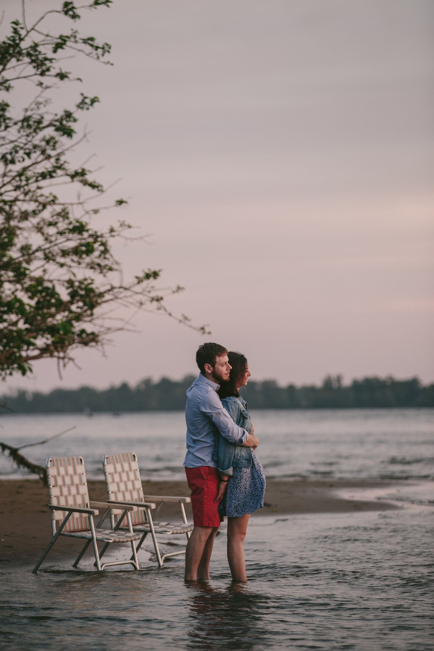 A romantic couple stands in shallow water at sunset with two empty chairs on the beach.