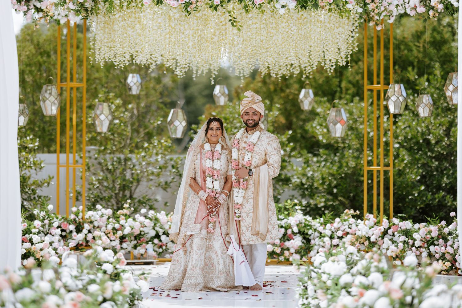 A couple in traditional Indian wedding attire sits on an ornate bench surrounded by white flowers in a garden setting.