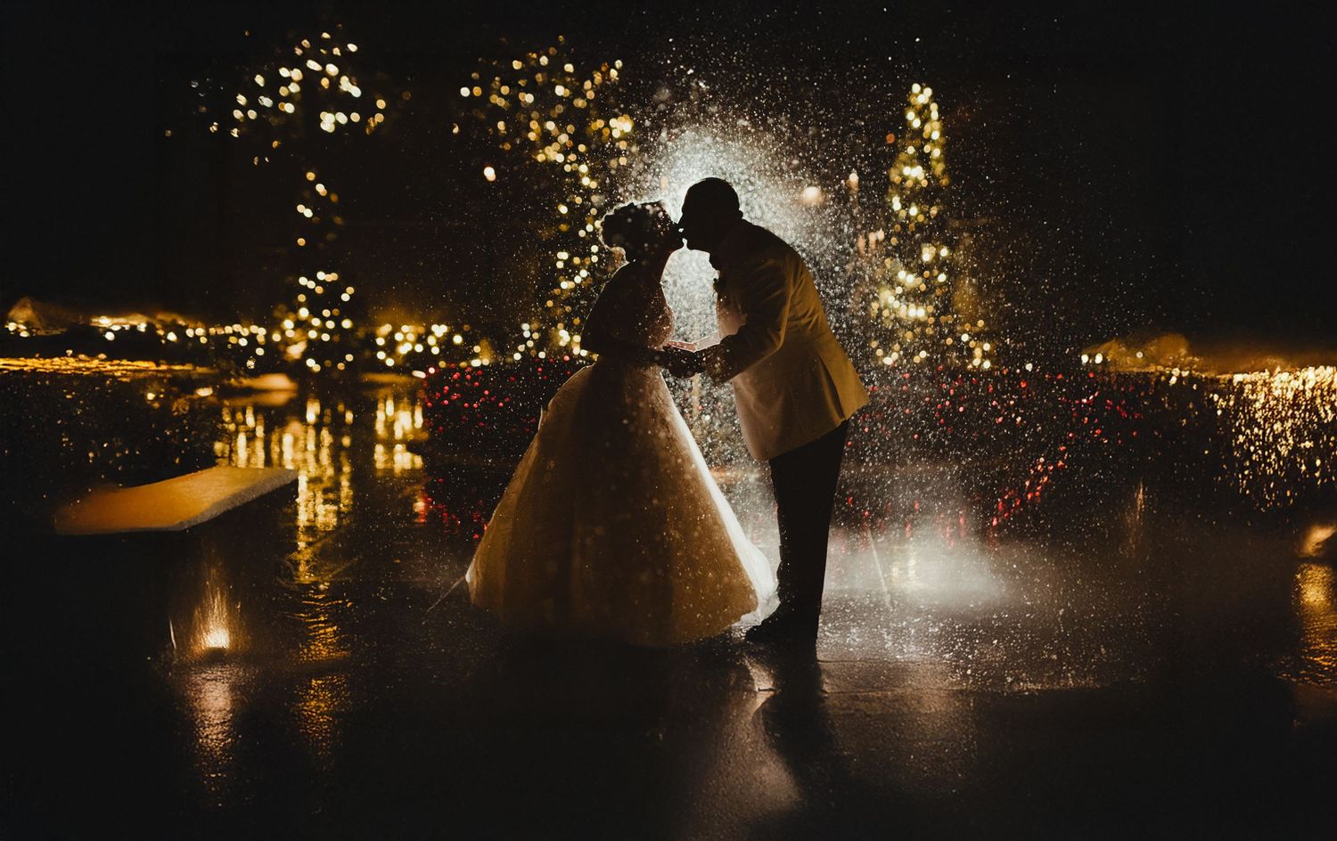 Silhouetted figures dance and celebrate amid sparkling fireworks and lights reflecting on wet ground at night.