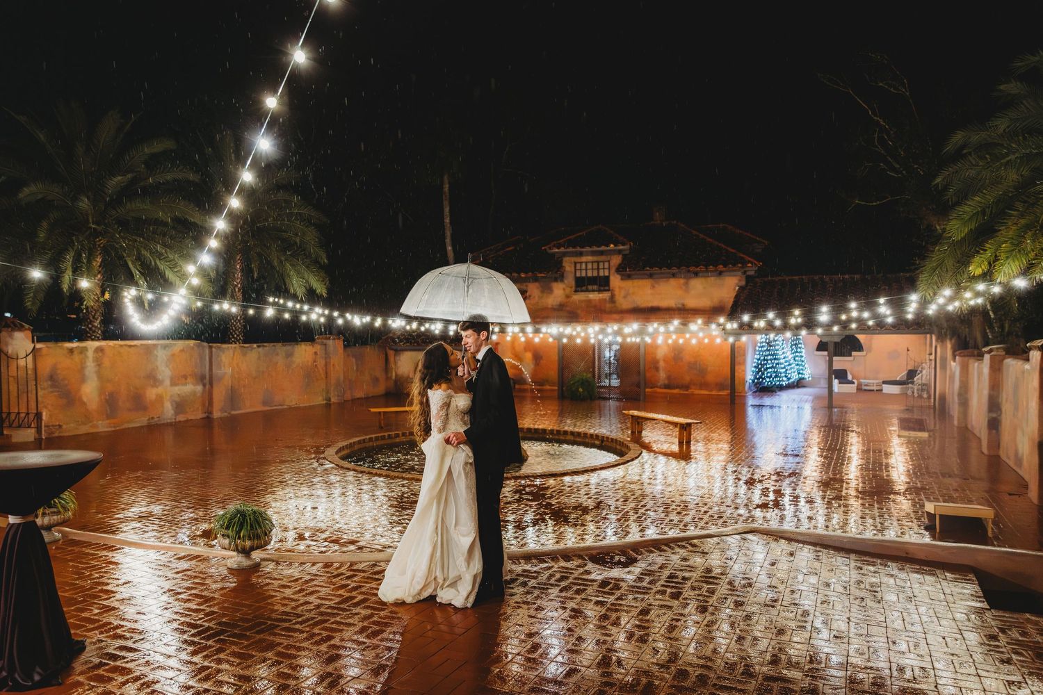 String lights illuminate an outdoor wedding reception venue with rows of chairs and tables under a starry night sky.