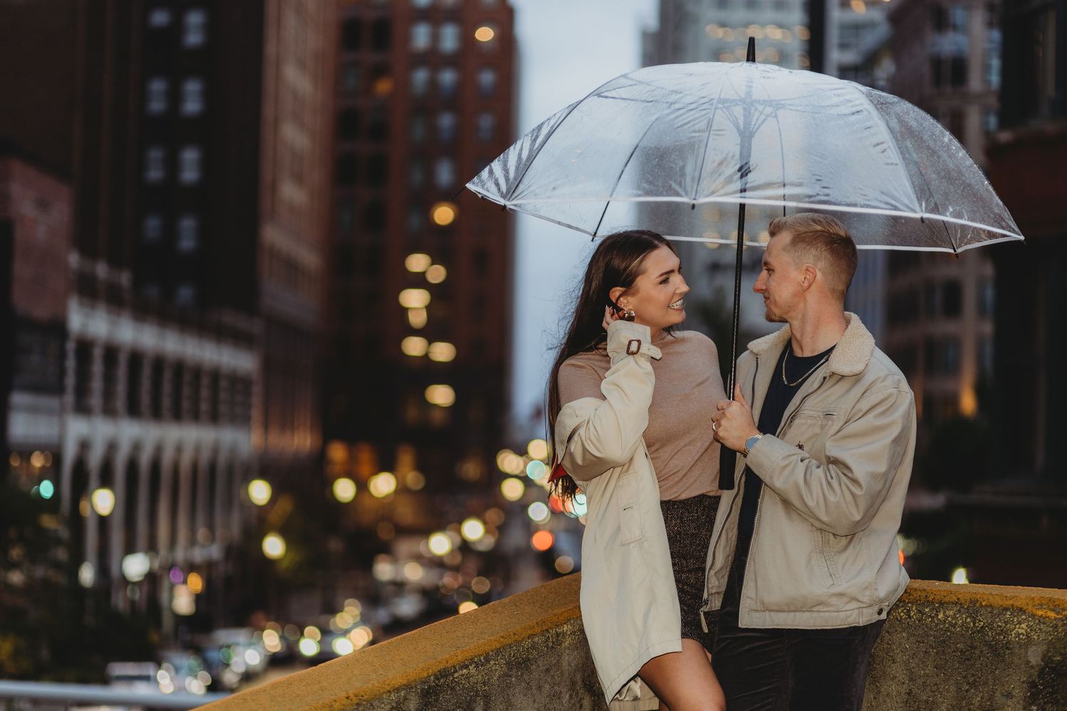 A couple shares romantic moments under clear umbrellas in an urban setting during evening hours.