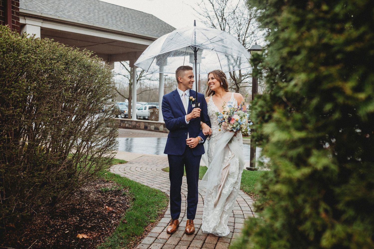 A couple in wedding attire walks along a stone path near a waterfront on an overcast day.