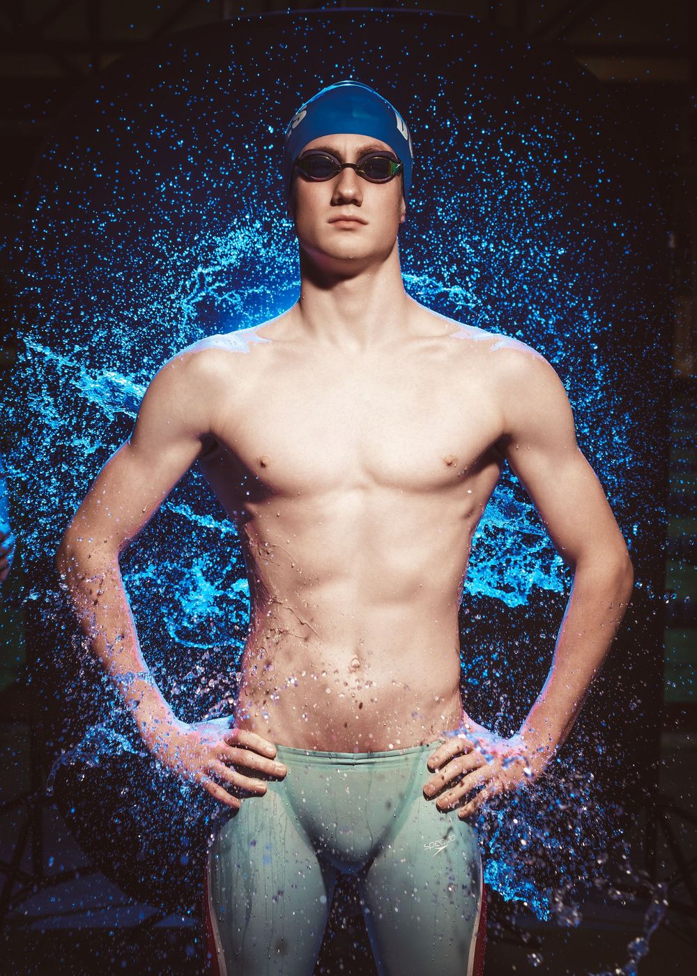 Athletic swimmer in blue swim cap and goggles stands against dark background with splashing water effects.