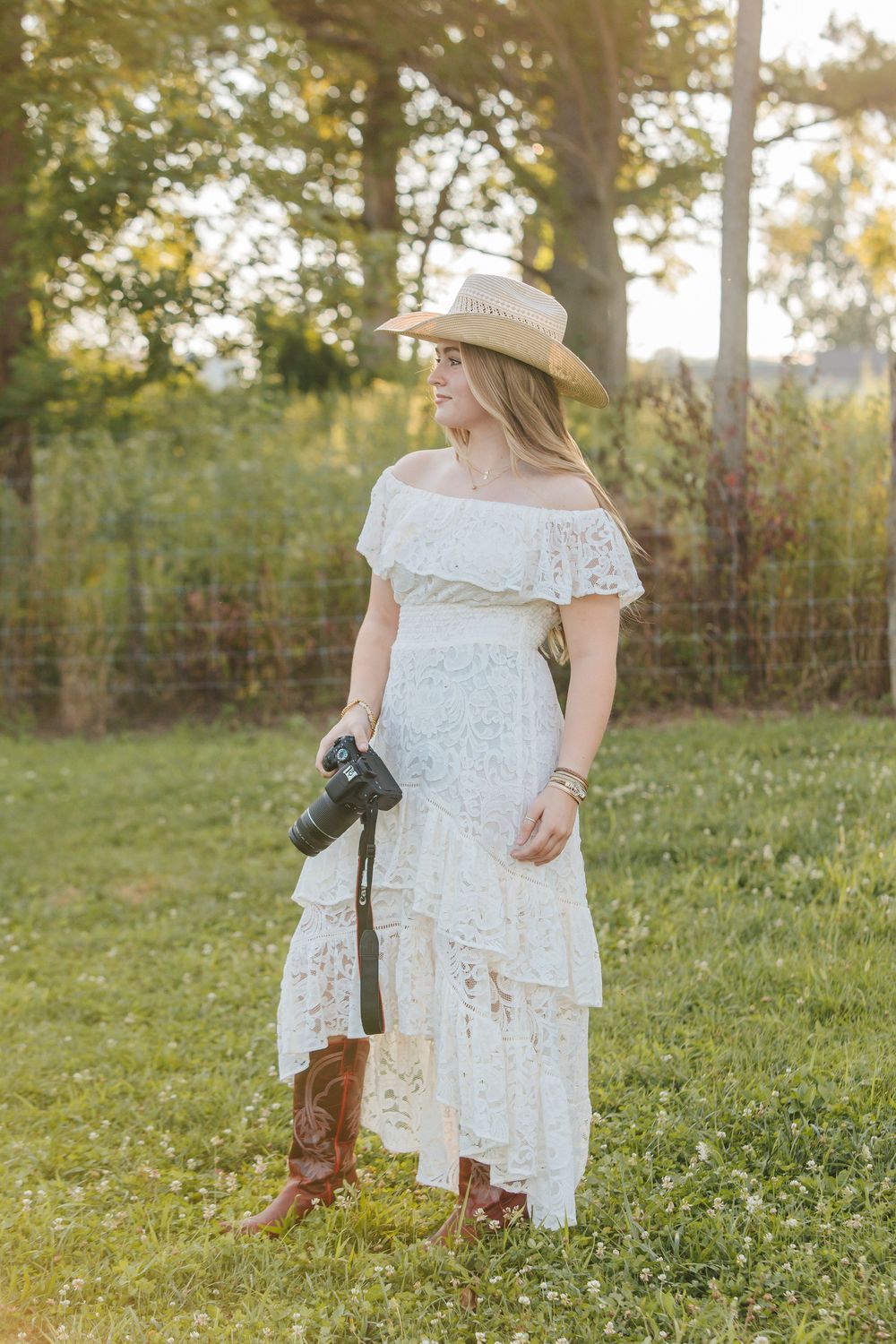 A photographer in a white lace off-shoulder dress and cowboy boots holds a camera in a sunlit meadow at golden hour.