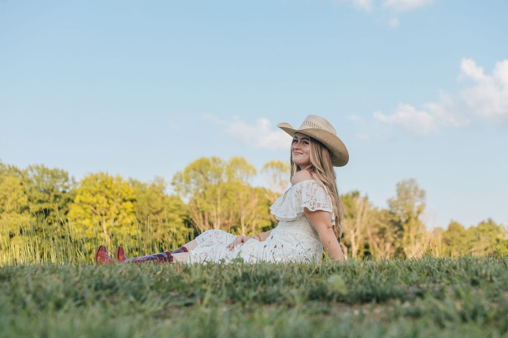 A cowgirl in a white lace dress sits on a hill posing for a picture.
