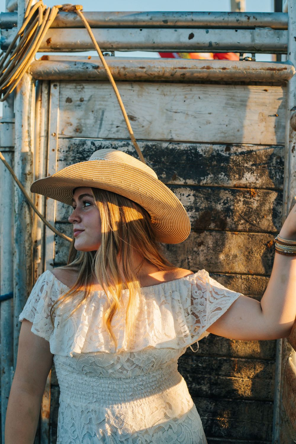 Person in white lace dress and straw hat poses against rodeo shoot in golden sunlight.