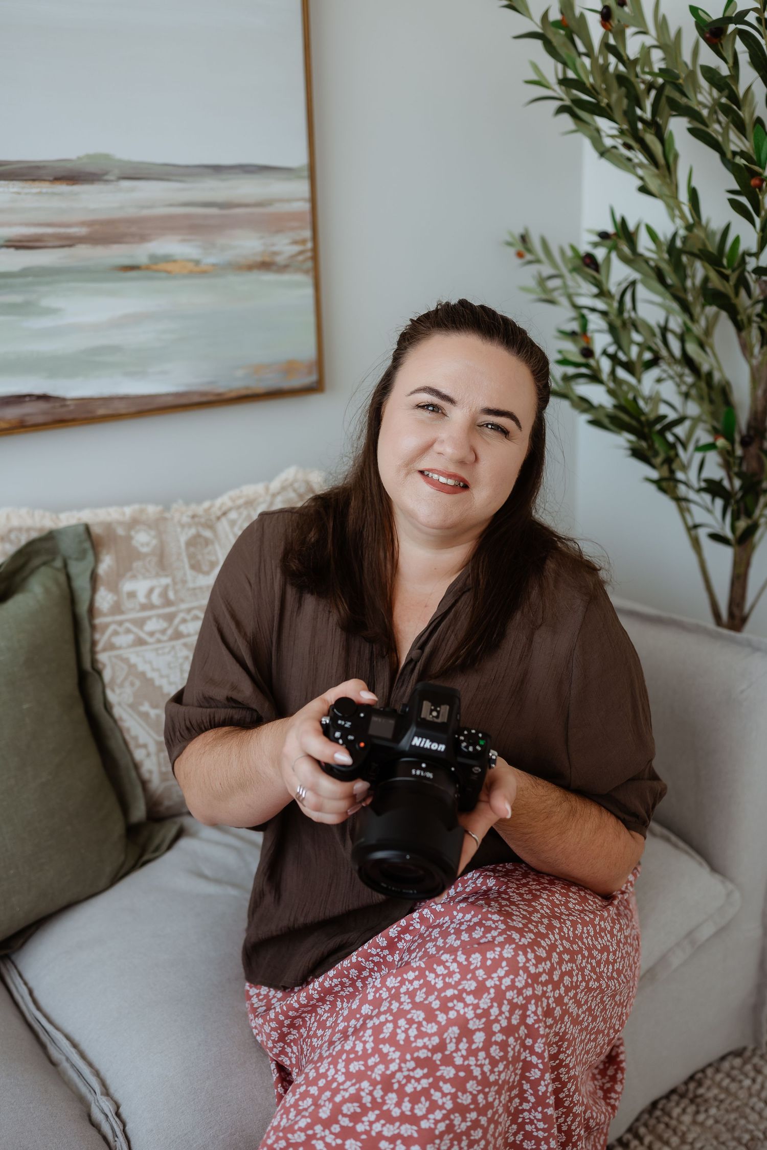 Photographer holding a camera while sitting on a couch with coastal artwork and plants in background at gladstone photography studio.