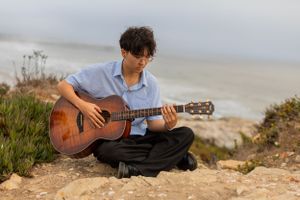 Guitarist performs on rocky oceanside cliff during overcast day.
