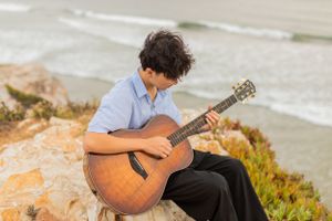 Musician playing acoustic guitar on coastal cliff overlooking ocean.