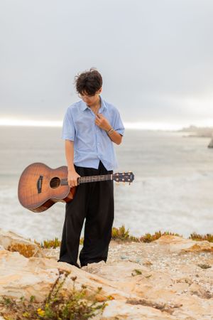 Musician with acoustic guitar standing on coastal cliffs at sunset in casual attire.