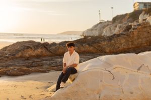 Young person in white shirt poses on coastal rocks during golden hour at beach.