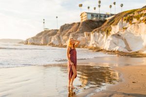 A person in a burgundy dress stands at the edge of ocean waves near dramatic coastal cliffs.
