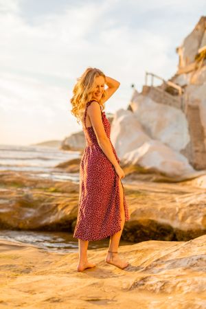 A woman in a red patterned dress poses on rocky coastal formations during sunset.