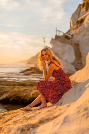 A person in a red dress sits on white coastal rocks during golden hour.