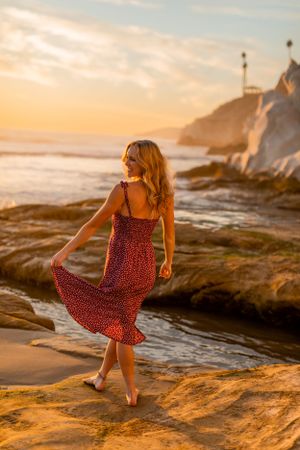 A figure in a flowing red dress twirls on rocky beach terrain at sunset.