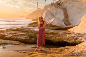 A person stands on rocky coastal formations in a red dress during sunset.
