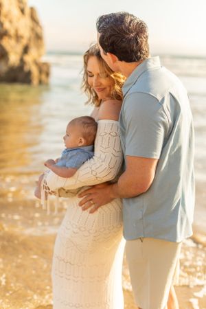 Couple holds baby while standing in golden sunlight on beach near rocky cliffs.