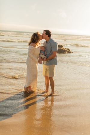 Romantic couple shares kiss while holding baby on sunset beach.