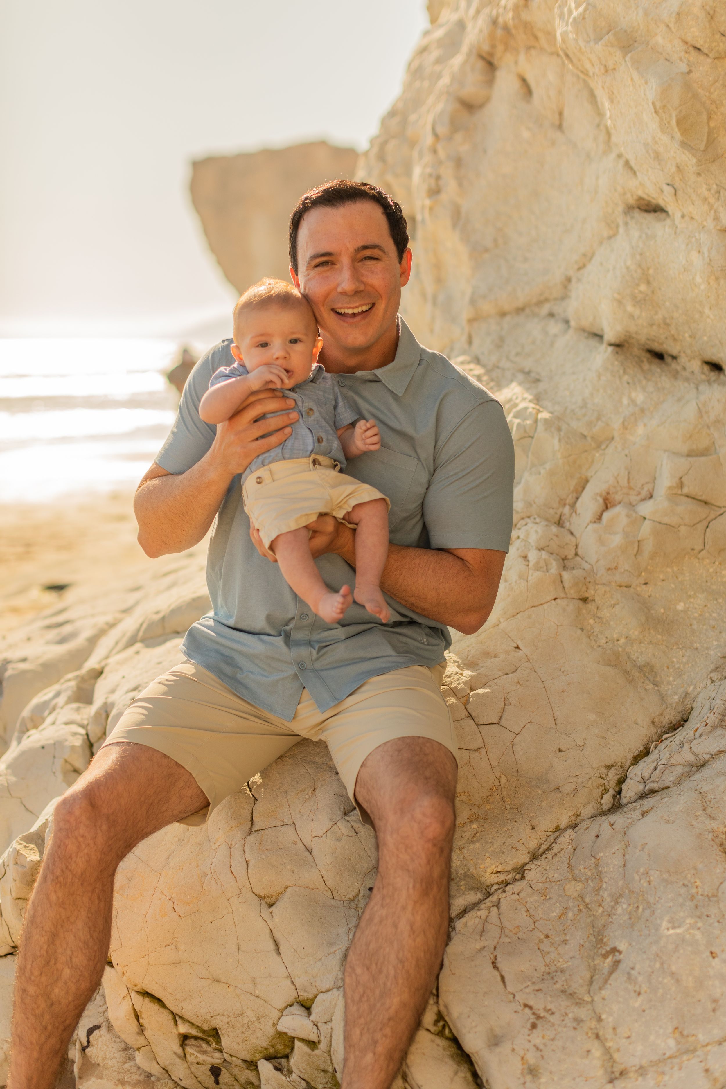 Parent and infant enjoy a golden sunset moment while sitting on coastal rocks at the beach.