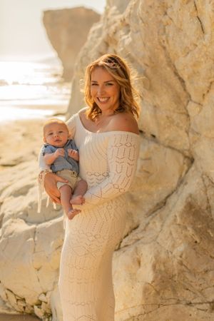 Woman in white lace dress holds baby against scenic beach cliffs during sunset photoshoot.