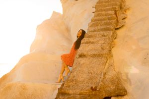 Person in orange dress climbs stone steps along dramatic coastal cliff face.