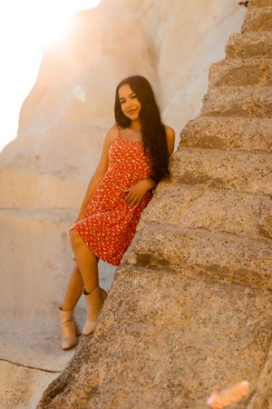 Woman in red floral dress poses on stone steps against sunlit limestone cliff.
