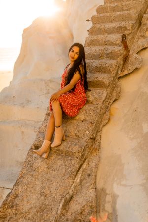 A woman in a red polka dot dress sits on weathered concrete steps against white cliffs at sunset.
