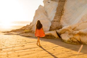 A figure in an orange dress walks along sandy beach by white coastal cliffs during golden hour.