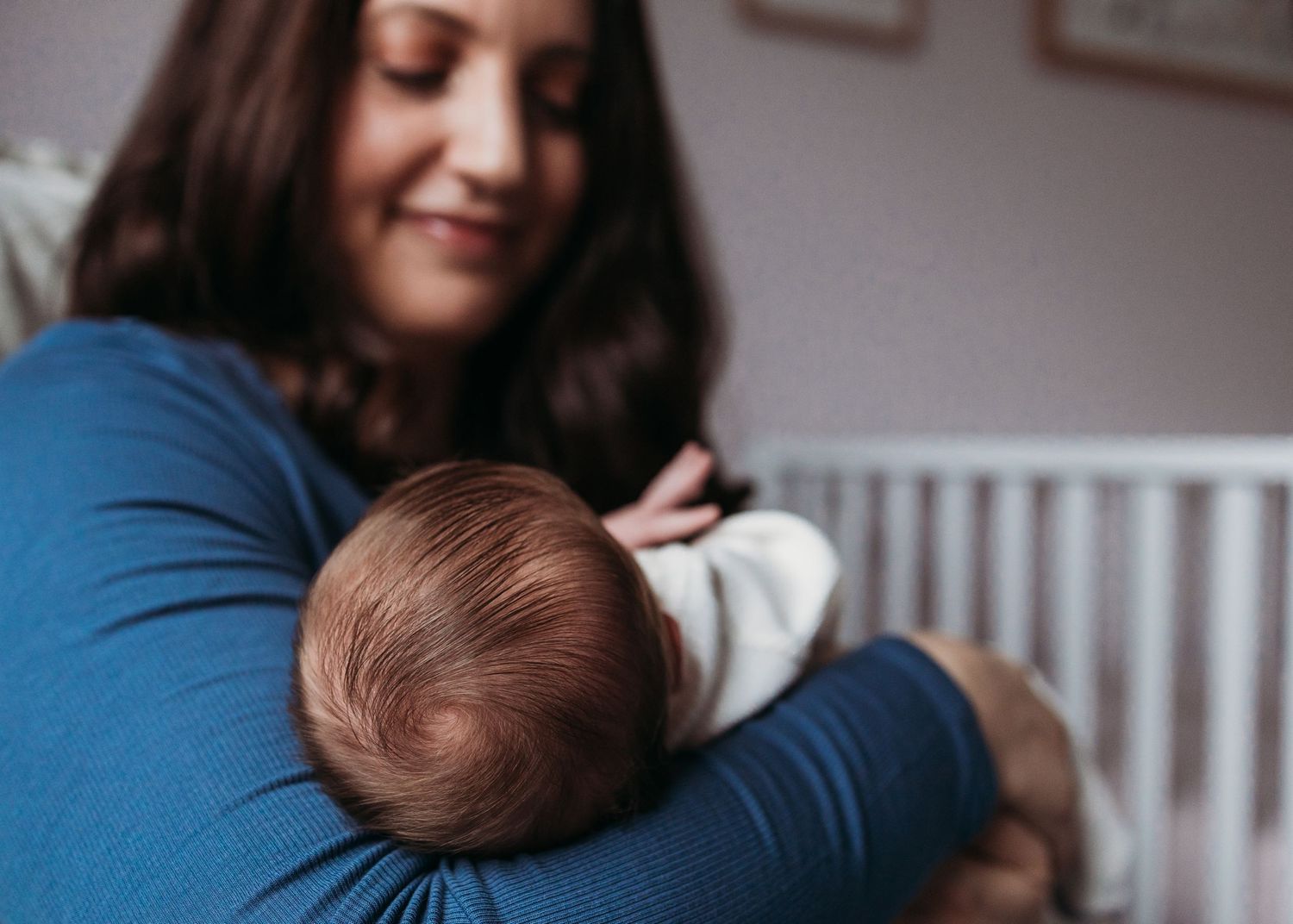 A parent in a blue sweater cradles a sleeping baby with brown hair against their chest in a soft, intimate moment.